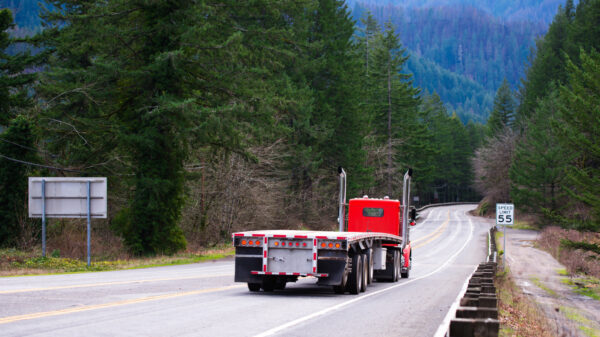 Red big rig semi truck tractor going with empty flat bed semi trailer on the awesome winding road in green forest in Columbia Gorge area alternate text for this image