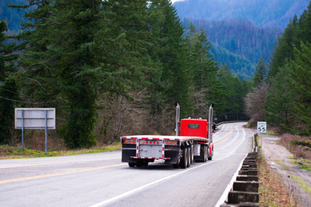 Red big rig semi truck tractor going with empty flat bed semi trailer on the awesome winding road in green forest in Columbia Gorge area alternate text for this image