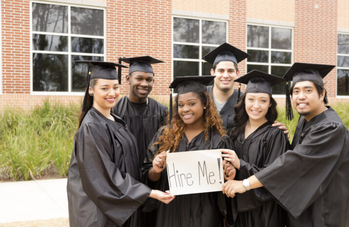 Six multi-ethnic graduates hold 'Hire Me' sign after college graduation. School building background. Employment issues concept.