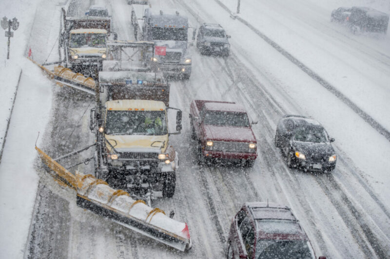 Two snow plows are clearing the road for vehicles in a winter storm.