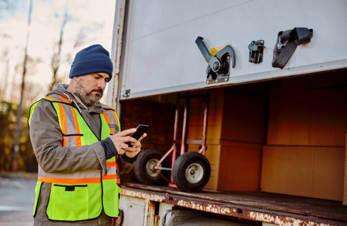 Mid adult courier using mobile phone while closing his truck and preparing for delivery.