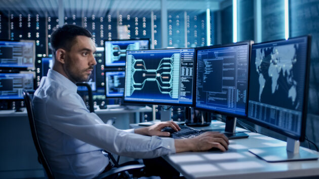 System Security Specialist Working at System Control Center. Room is Full of Screens Displaying Various Information. alternate text for this image
