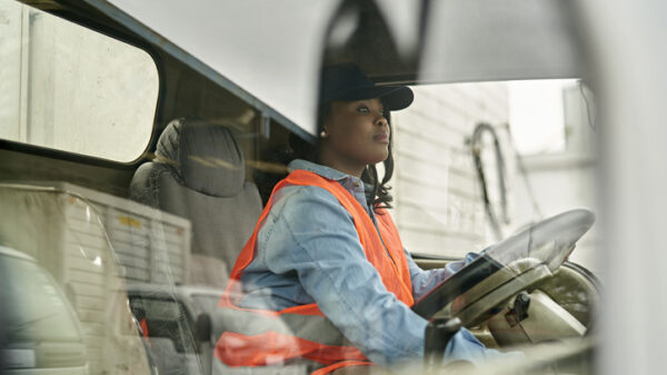Personal perspective of mid 20s woman in casual clothing, cap, and reflective vest sitting in driver’s seat, looking straight ahead, ready to begin road trip.