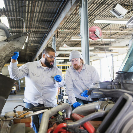 Two multi-ethnic mechanics repairing a semi-truck. They are looking under the hood of the truck, examining the engine. The African-American man is in his 30s and his coworker is in his 40s.