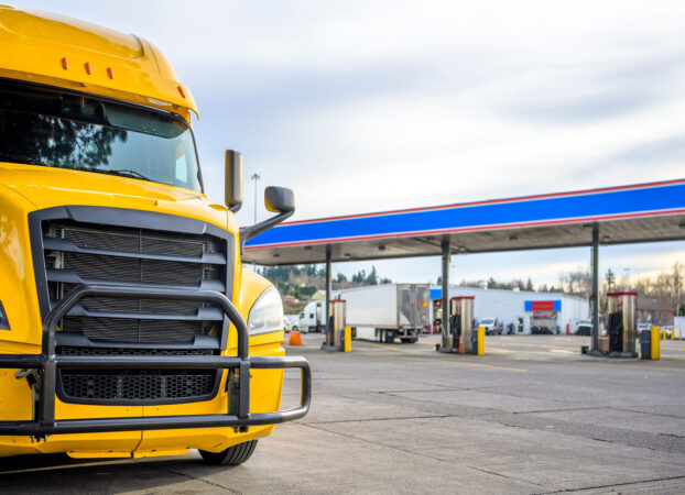 Big rig yellow long haul industrial diesel semi truck with grille guard standing in row with another semi trucks and semi trailers on truck stop parking lot with fuel station for refueling