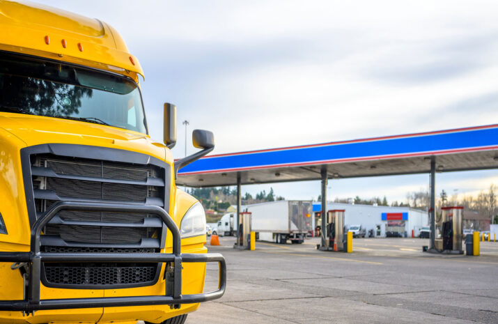 Big rig yellow long haul industrial diesel semi truck with grille guard standing in row with another semi trucks and semi trailers on truck stop parking lot with fuel station for refueling