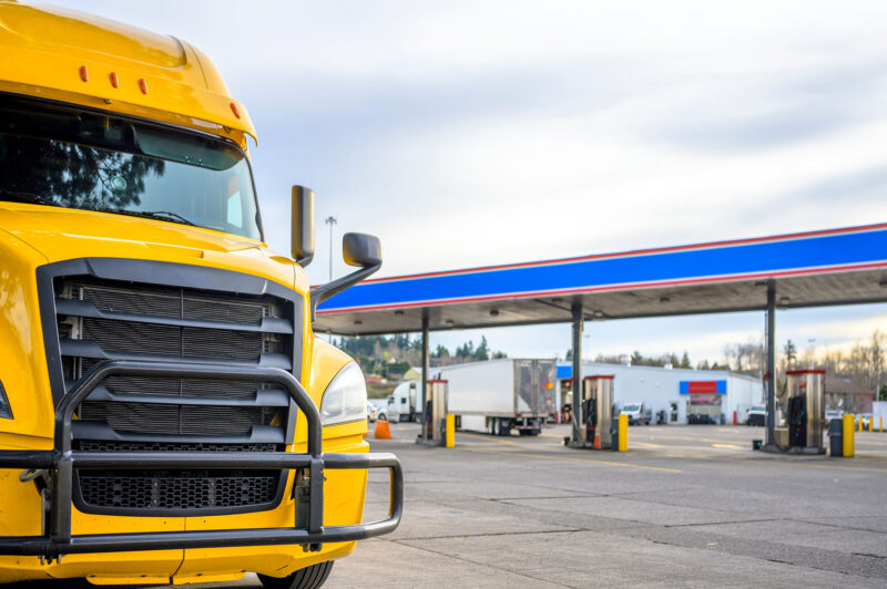 Big rig yellow long haul industrial diesel semi truck with grille guard standing in row with another semi trucks and semi trailers on truck stop parking lot with fuel station for refueling