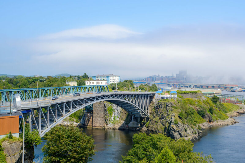 Reversing Falls bridge in Saint John, NB, Canada, seen from Wolastoq Park. Fog creeps over the city center in the distance. Sunny day.