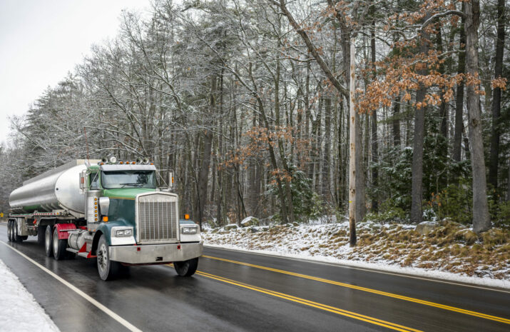 Industrial green day cab classic bonnet big rig semi truck transporting liquid cargo in long oval cistern tank semi trailer running on the wet winter road with snow on the sides and snowy forest