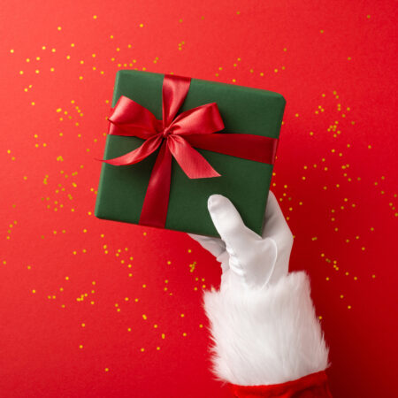 Santa Claus hand holding a green gift box with a red ribbon on a festive square red background, conveying holiday spirit and celebration alternate text for this image