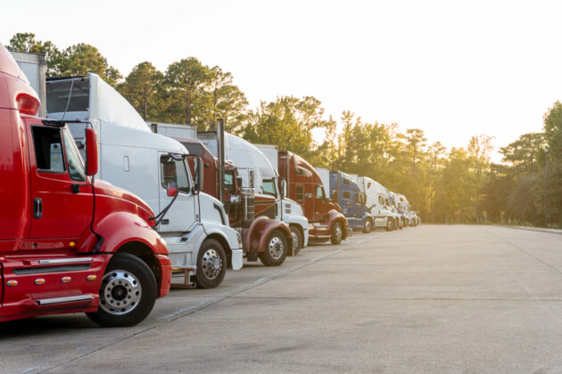 Row of semi trucks parked in lot, under a clear sky.