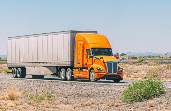 Bright orange truck hauling a long trailer on a rural highway through a dry, open landscape.
