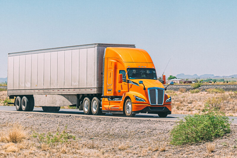 Bright orange truck hauling a long trailer on a rural highway through a dry, open landscape.