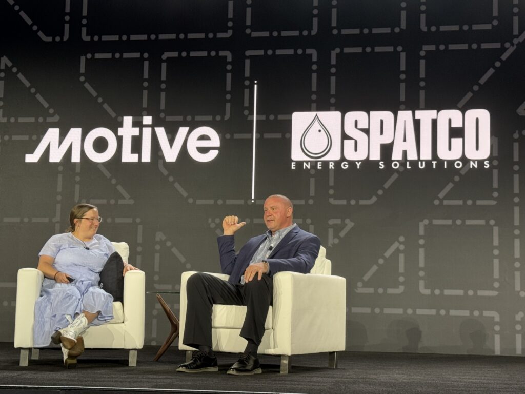 Two people sitting on stage in chairs during a conference, Motive and Spatco logos on background