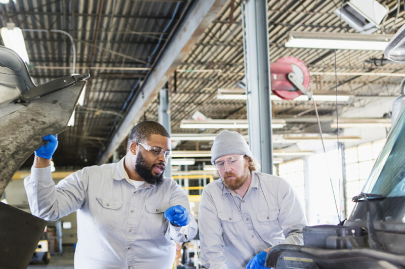 Two multi-ethnic mechanics repairing a semi-truck. They are looking under the hood of the truck, examining the engine. The African-American man is in his 30s and his coworker is in his 40s.