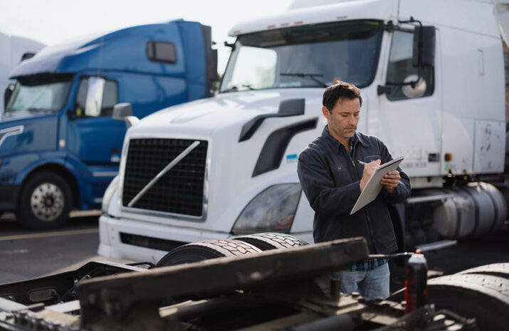 Truck driver writing notes while inspecting a semi-truck trailer in a parking lot, with other trucks in the background